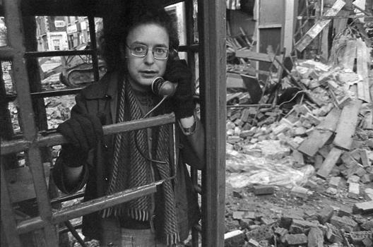 Stewart Parker standing in a phone booth in a street covered in rubble from a bombing.