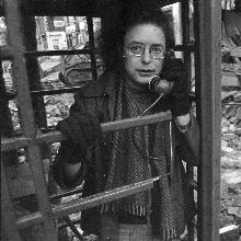 Stewart Parker standing in a phone booth in a street covered in rubble from a bombing.